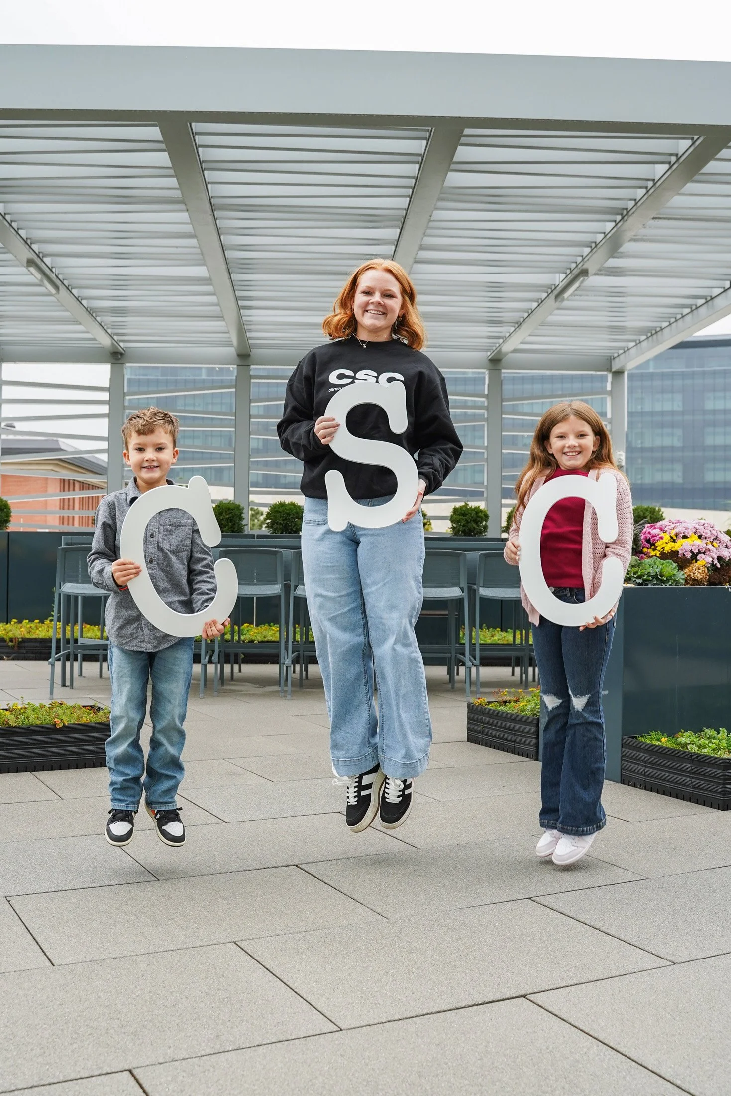 Three children and one young adult holding large white letters spelling 'CSC' in an outdoor urban setting with a modern building and greenery in the background.