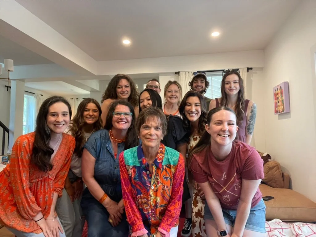 Group of people gathered inside a living room, smiling for a group photo. There are men and women of various ages, some with long hair, casual clothing, and a woman in colorful clothing in the front center.