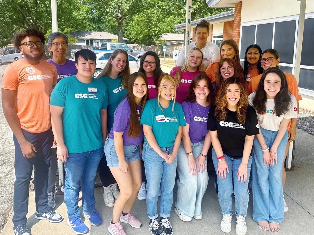 Group of young people and adults standing outdoors on a sunny day, some wearing CSC shirts, with a building and parked cars in the background.