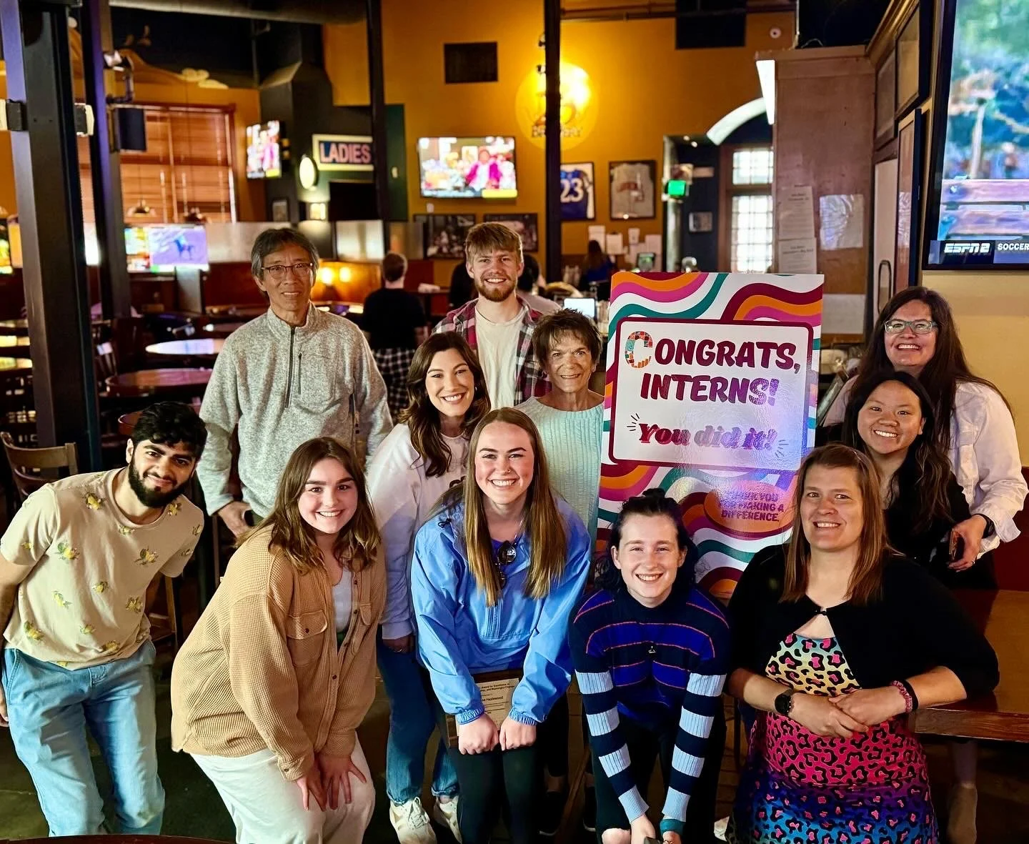 A group of people posing together indoors in a restaurant or bar, celebrating interns with a sign that reads, 'Congrats, Interns! You did it!'