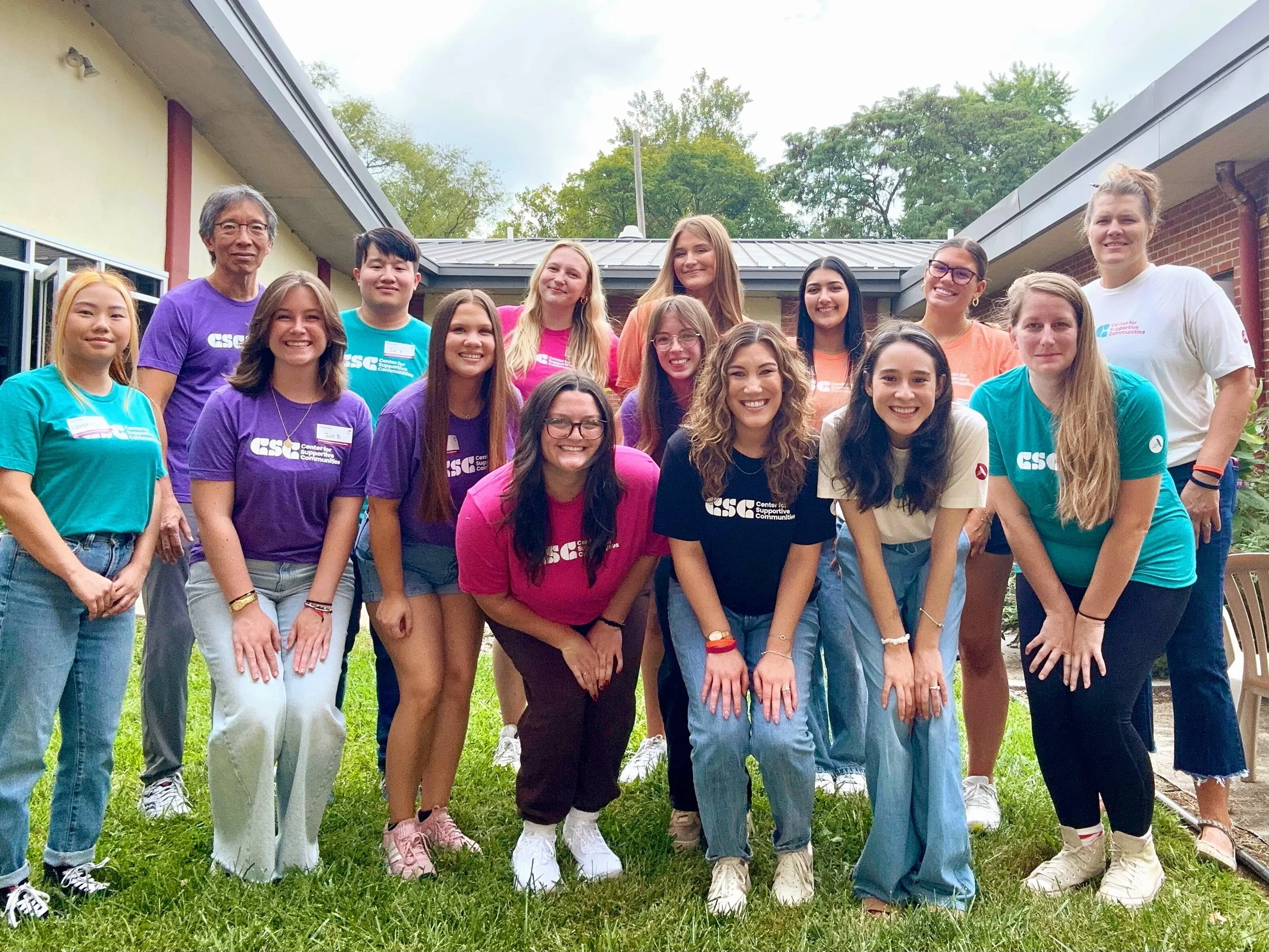 Group of young diverse people standing together outdoors, smiling for a photo, with some wearing colorful t-shirts that read 'Center for Supportive Communities'.