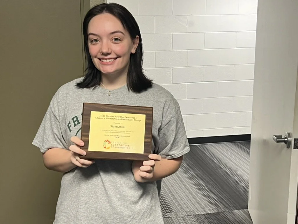 A young woman with black hair and a gray t-shirt smiling and holding a plaque. The plaque has text that mentions an award for excellence in advocacy, mentorship, and meaningful change.