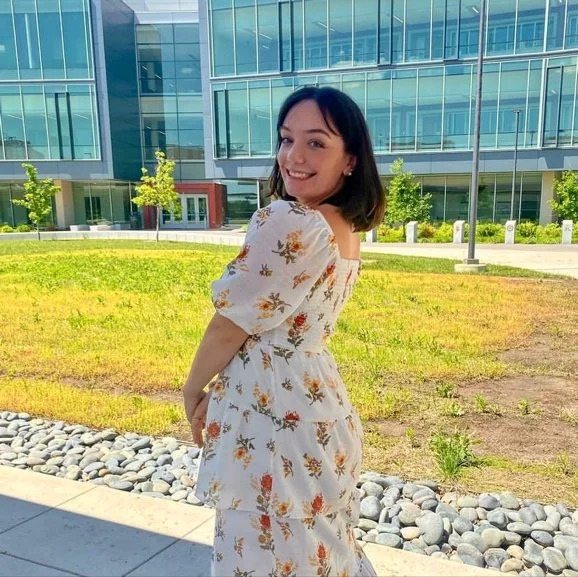 Smiling young woman with shoulder-length dark hair, wearing a white floral dress, standing outside in front of a modern glass building and green lawn.