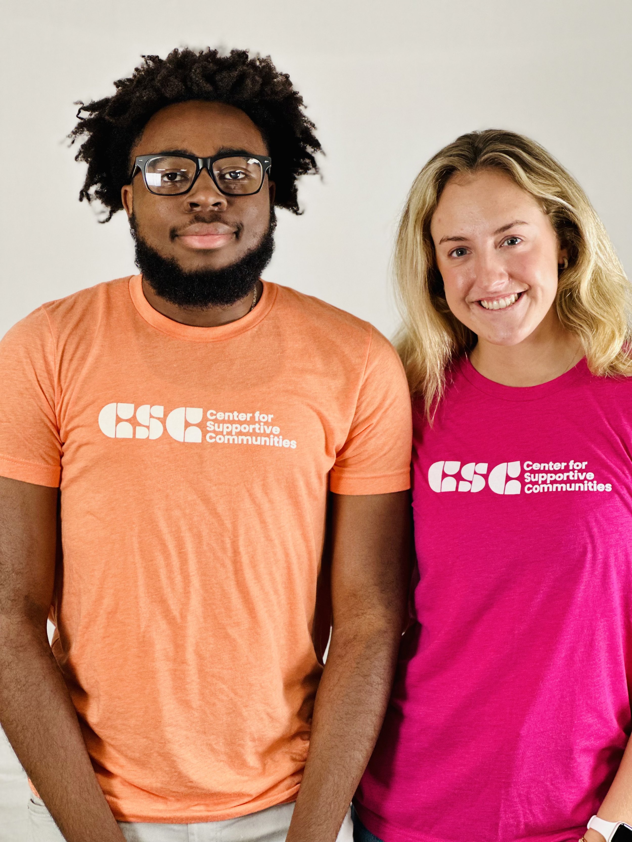 Two people standing side by side, wearing orange and pink shirts with the logo and text 'Center for Supportive Communities' on them.