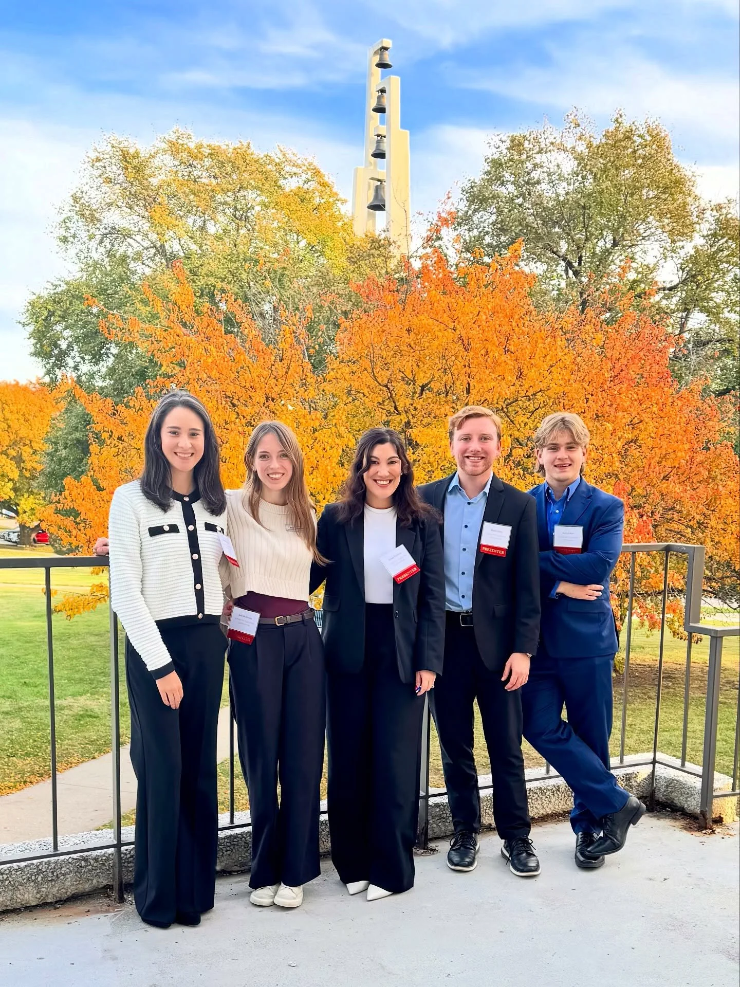 Group of five young adults standing outdoors on a concrete path with a fence, autumn trees in background showing orange and green leaves, and a bell tower in the distance. They are smiling, dressed in business casual to formal attire, and wearing name tags.