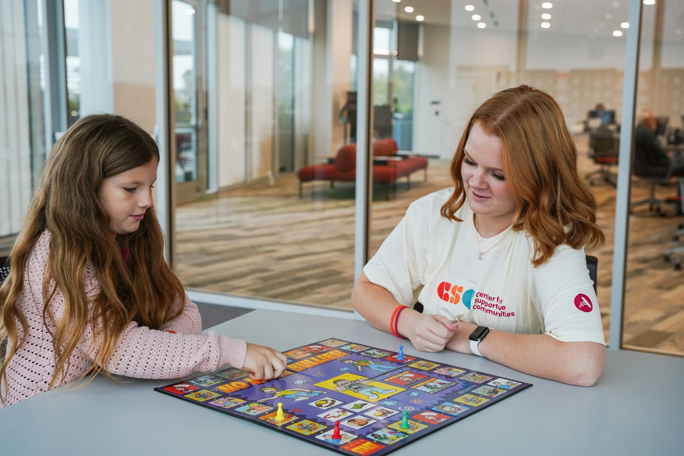 A girl and a woman playing a board game at a table indoors, with glass walls and a modern interior in the background.