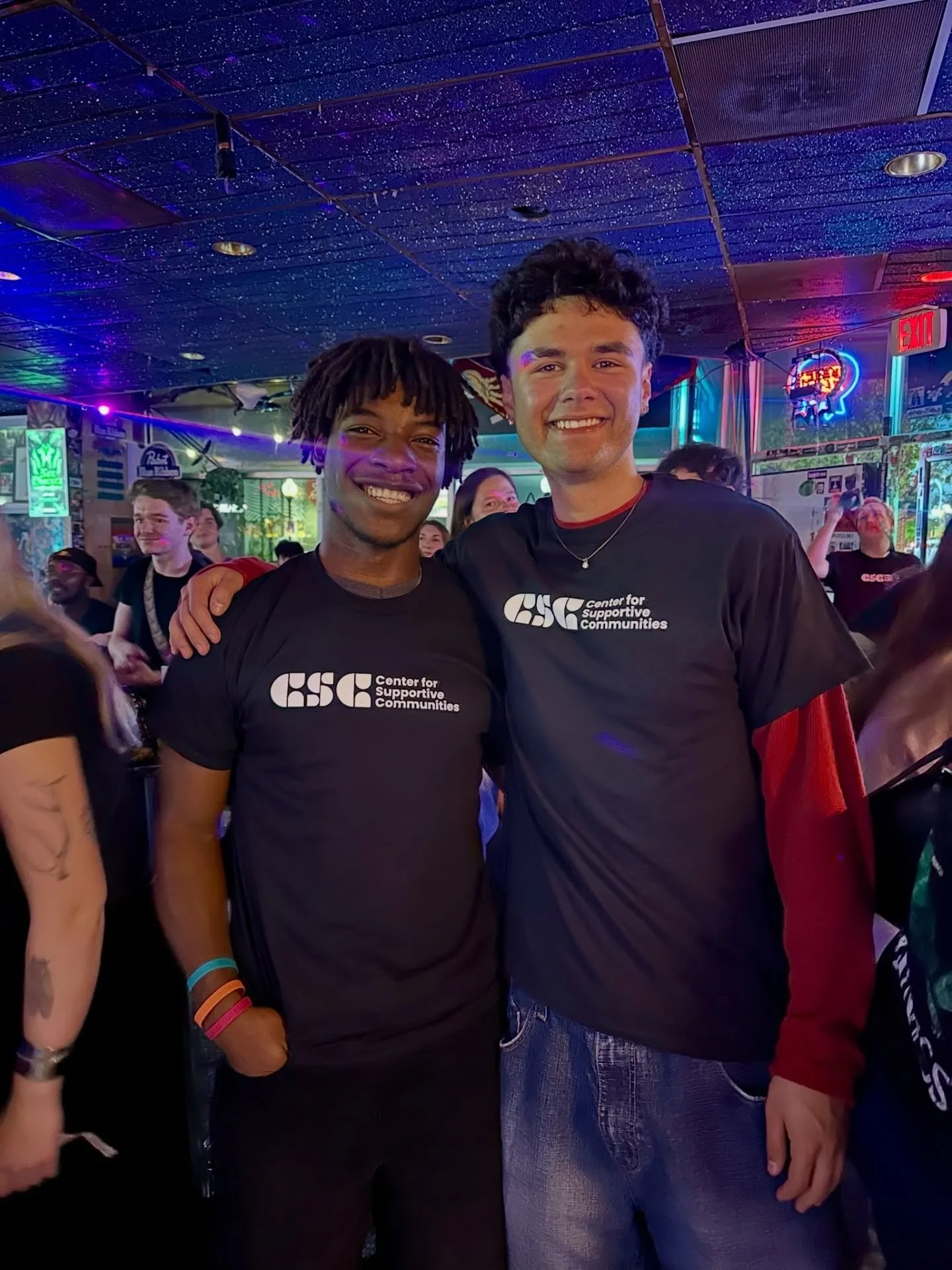 Two young men smiling and posing together at a lively indoor event with colorful neon signs and a starry ceiling. They are wearing black shirts with white text that reads 'CSC Center for Supportive Communities.' One has dreadlocks, and the other has curly hair.