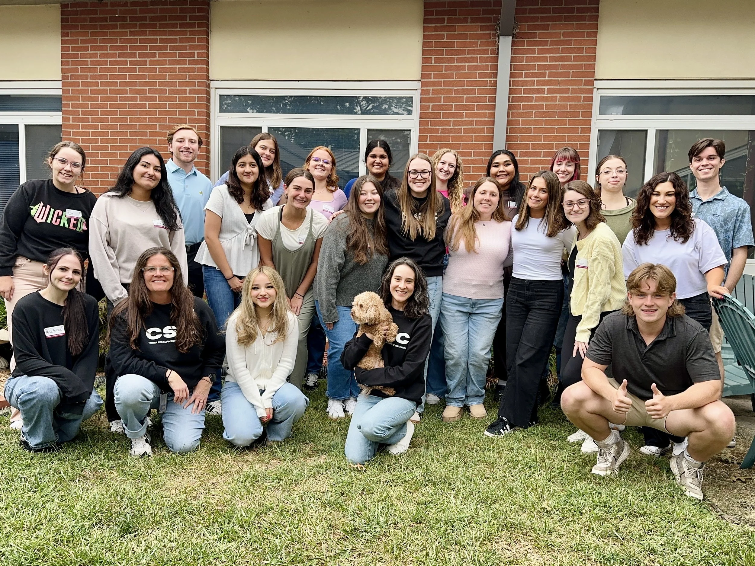 Group of diverse young adults posing for a photo outdoors in front of a brick building, some kneeling in front and some standing behind, with a green lawn and a dog in the front.