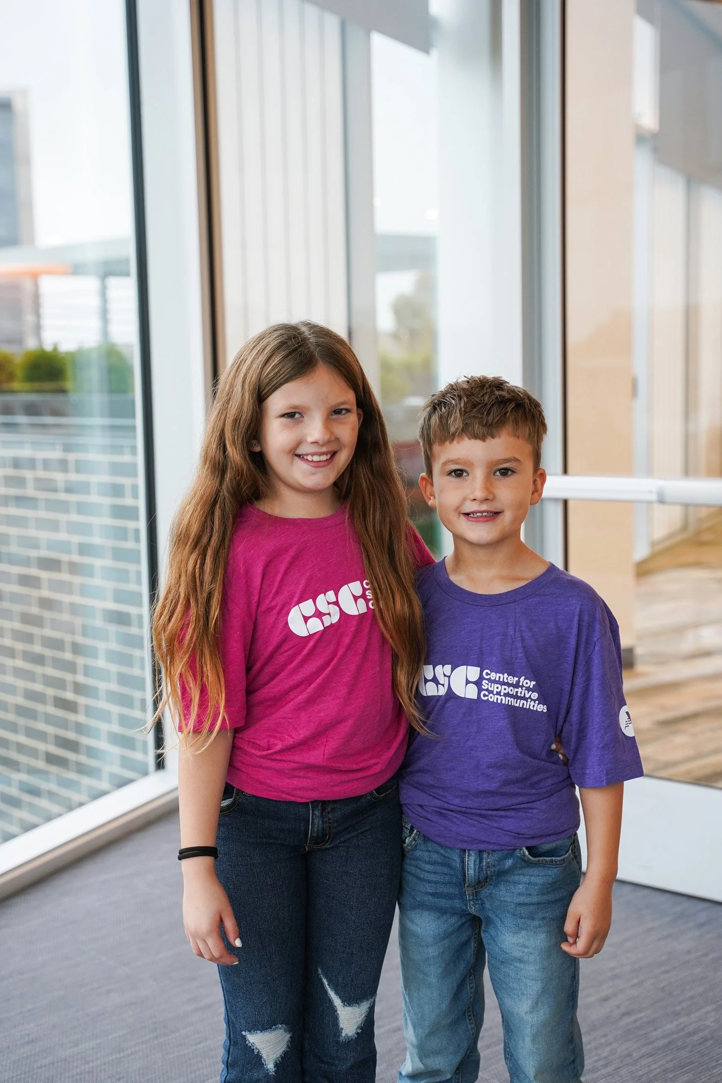 Two children, a girl with long brown hair and a boy with short brown hair, standing in a brightly lit indoor space near large windows, both wearing T-shirts with the 'Center for Supportive Communities' logo. The girl is smiling and wearing a pink T-shirt, while the boy is smiling and wearing a purple T-shirt. The girl has ripped jeans and a black bracelet, and they are standing close together.