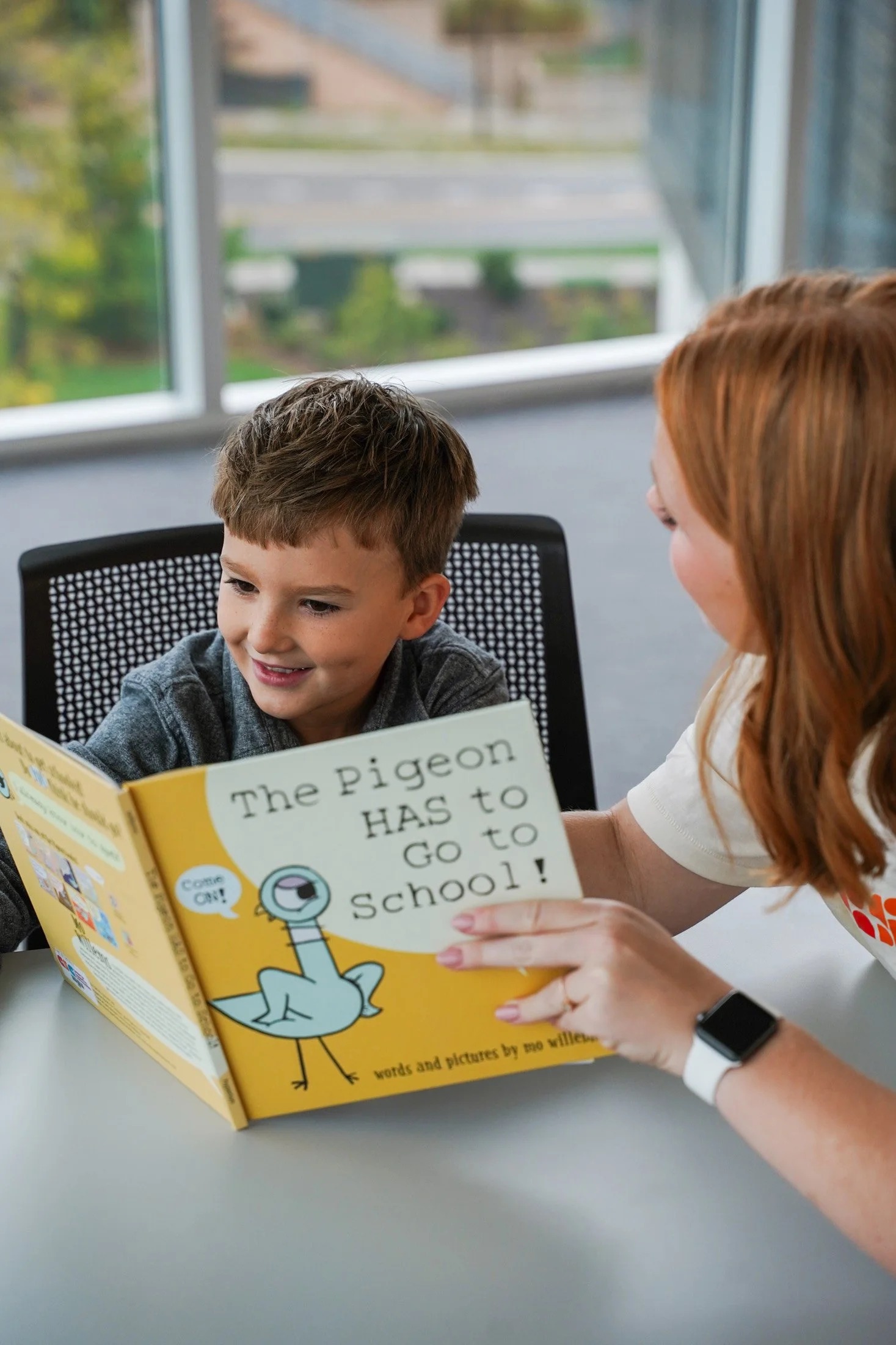 A young boy holding a children's book titled "The Pigeon HAS to Go to School!" while a woman, possibly his teacher or parent, helps him read. They are seated at a table in a room with large windows and an outdoor view.