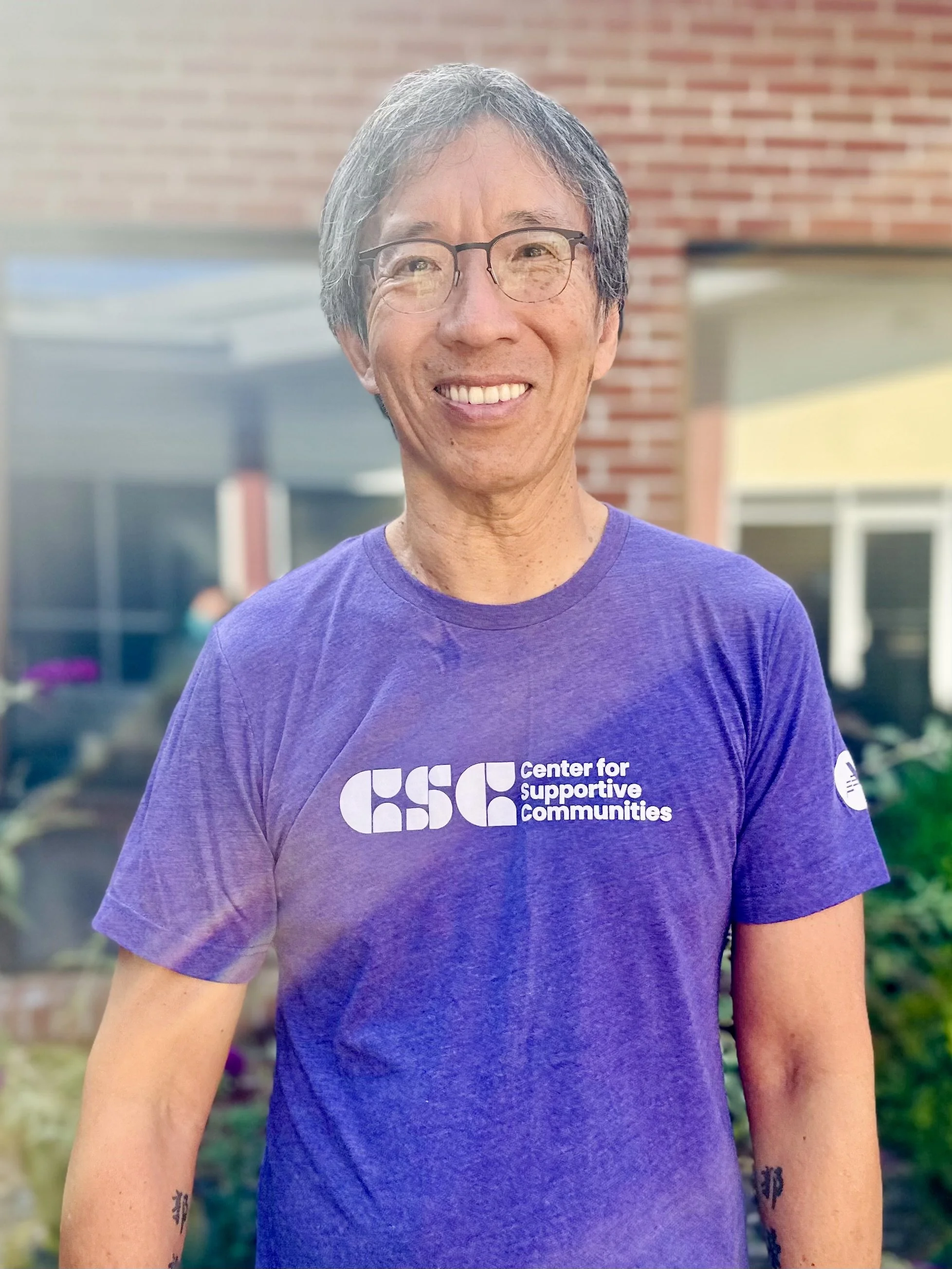 A smiling person with gray hair and glasses, wearing a purple T-shirt with a logo and text for the Center for Supportive Communities, standing outdoors in front of a brick building.