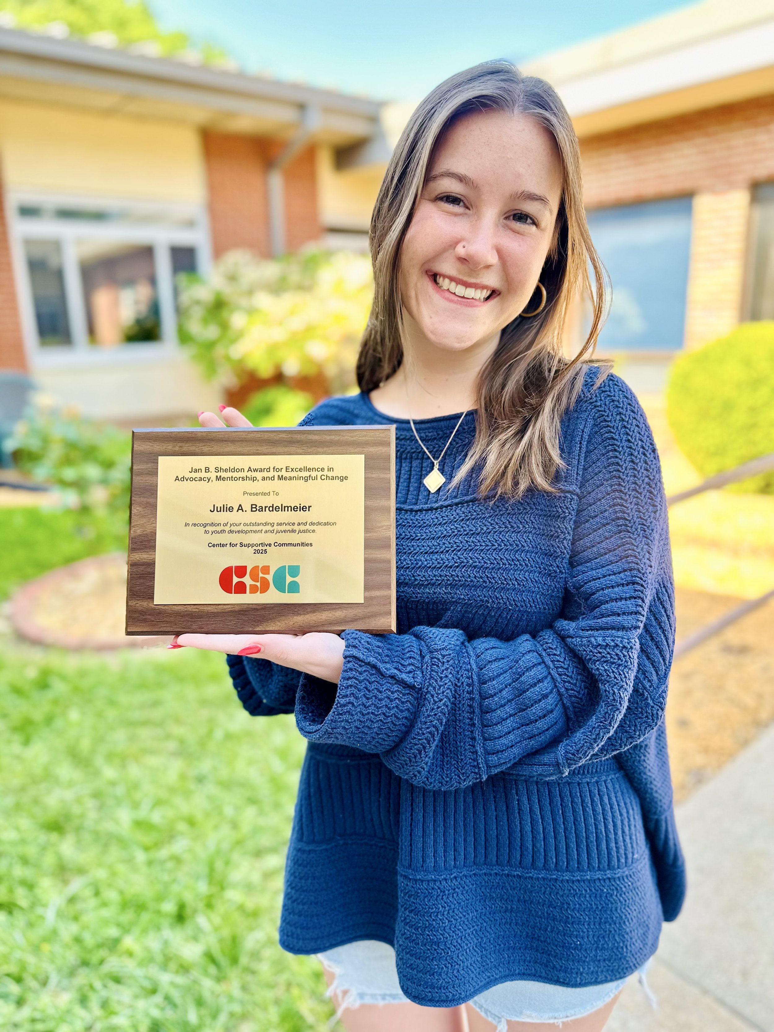 A woman with light brown hair smiling, holding a wooden award plaque outdoors on a sunny day. She is wearing a blue sweater and white shorts.