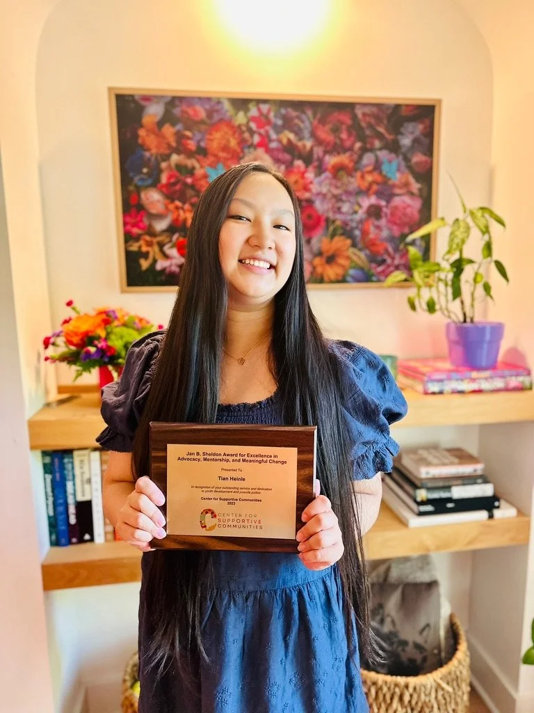 Young woman smiling and holding an award plaque in a room with colorful floral artwork, bookshelves, and houseplants.