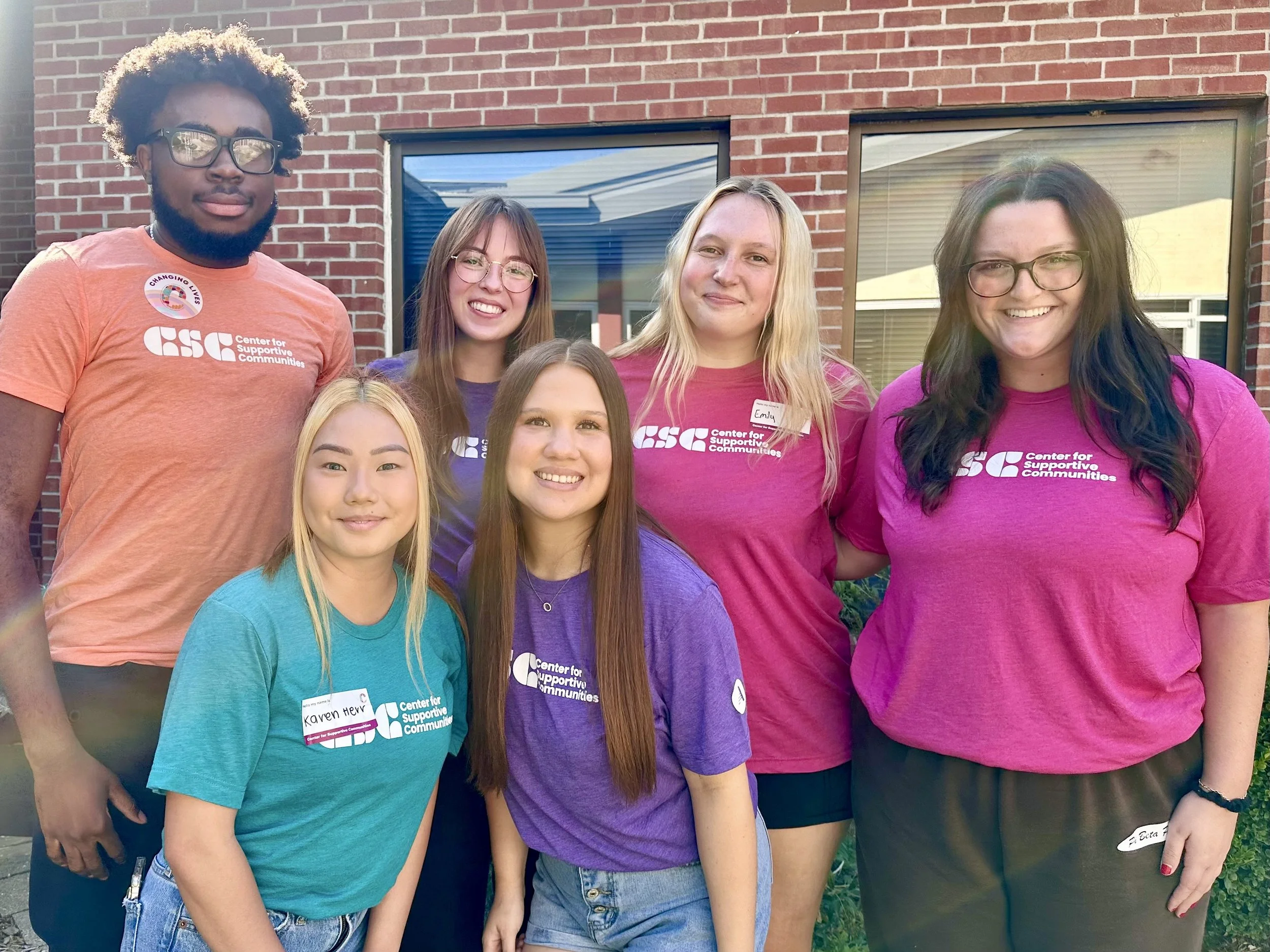 Group of six young women standing outside in front of a red brick building, smiling at the camera. They are wearing colorful t-shirts with the logo of the Center for Supportive Communities, some with name tags.