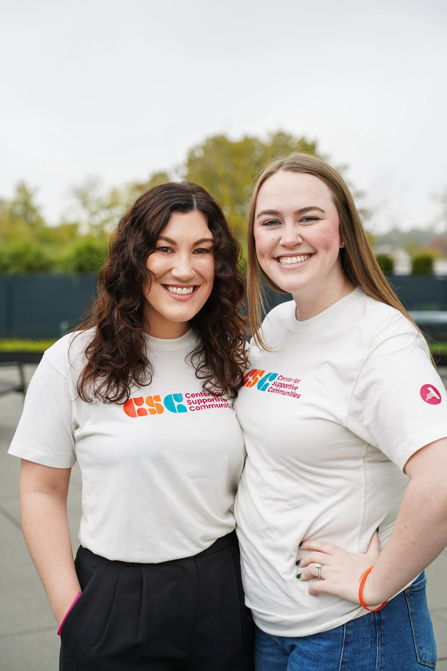 Two smiling women standing outdoors, wearing white t-shirts with colorful logos, with background of trees and a cloudy sky.