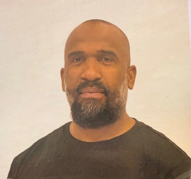 A man with a beard and short hair wearing a black T-shirt standing against a plain white background.