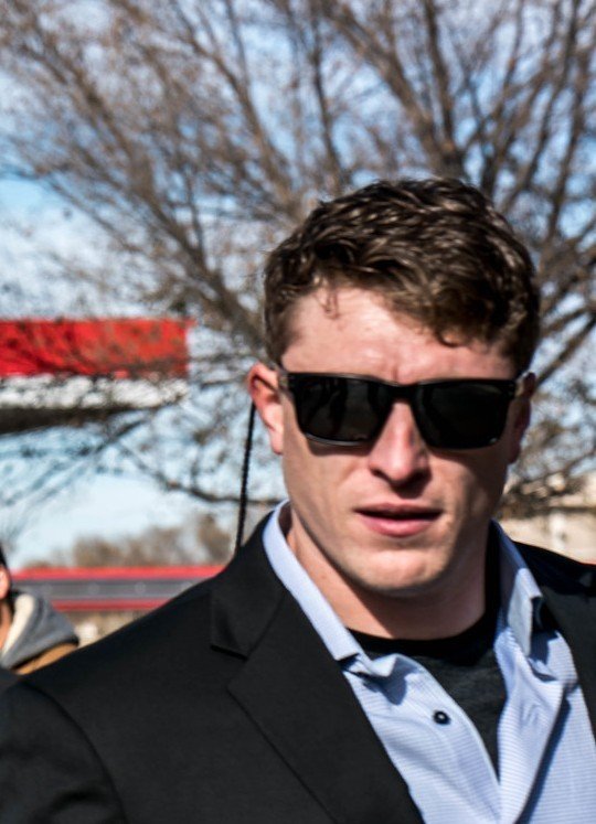 Young man with dark curly hair wearing sunglasses, a black blazer, and a light blue dress shirt outdoors on a sunny day, with leafless trees in the background.
