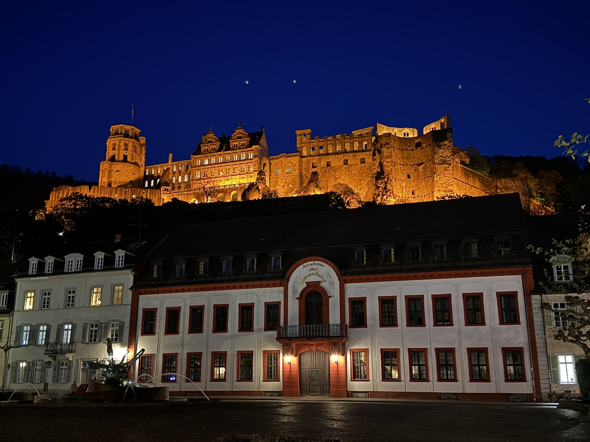 Night view of a castle illuminated in orange lights on a hill with a dark blue sky and stars above. Below, a row of buildings with lit windows and a water fountain in front.