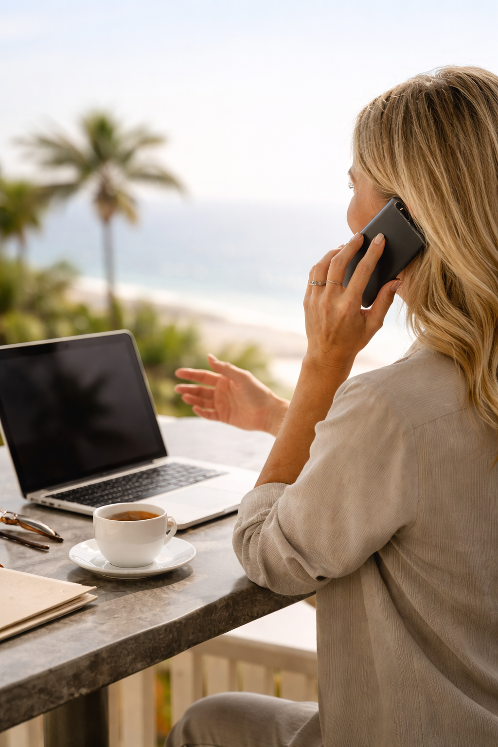 Woman with blonde hair talking on her cellphone at a seaside outdoor workspace, with a laptop, cup of coffee, and sunglasses on the table, overlooking palm trees and the ocean.