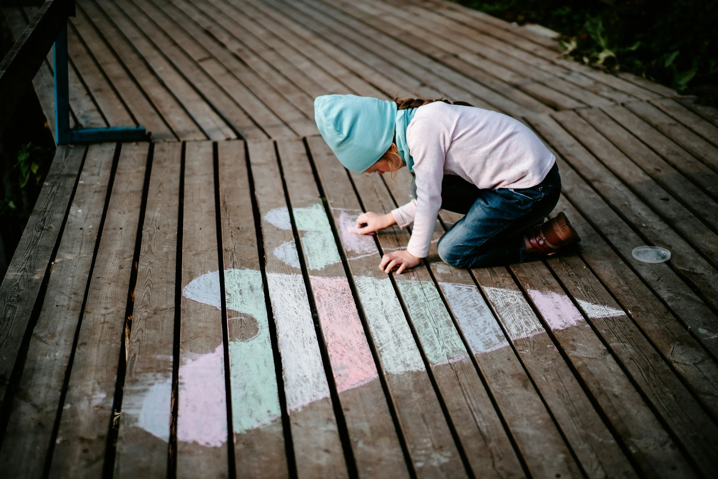 A young girl wearing a blue beanie, pastel pink sweater, and jeans is kneeling on a wooden deck drawing with colorful chalk
