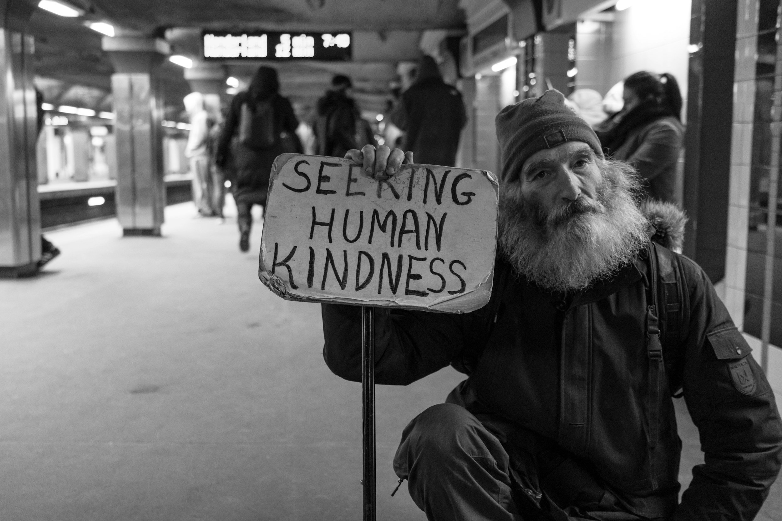 A bearded man wearing a beanie hat and dark jacket kneels at an underground subway station, holding a sign that reads 'Seeking Human Kindness'. The station is populated with a few other people in the background.