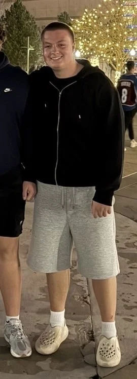 Young man with a big smile, wearing a black hoodie, gray shorts, white socks, and beige Crocs, standing outdoors at night with lit trees and people in the background.