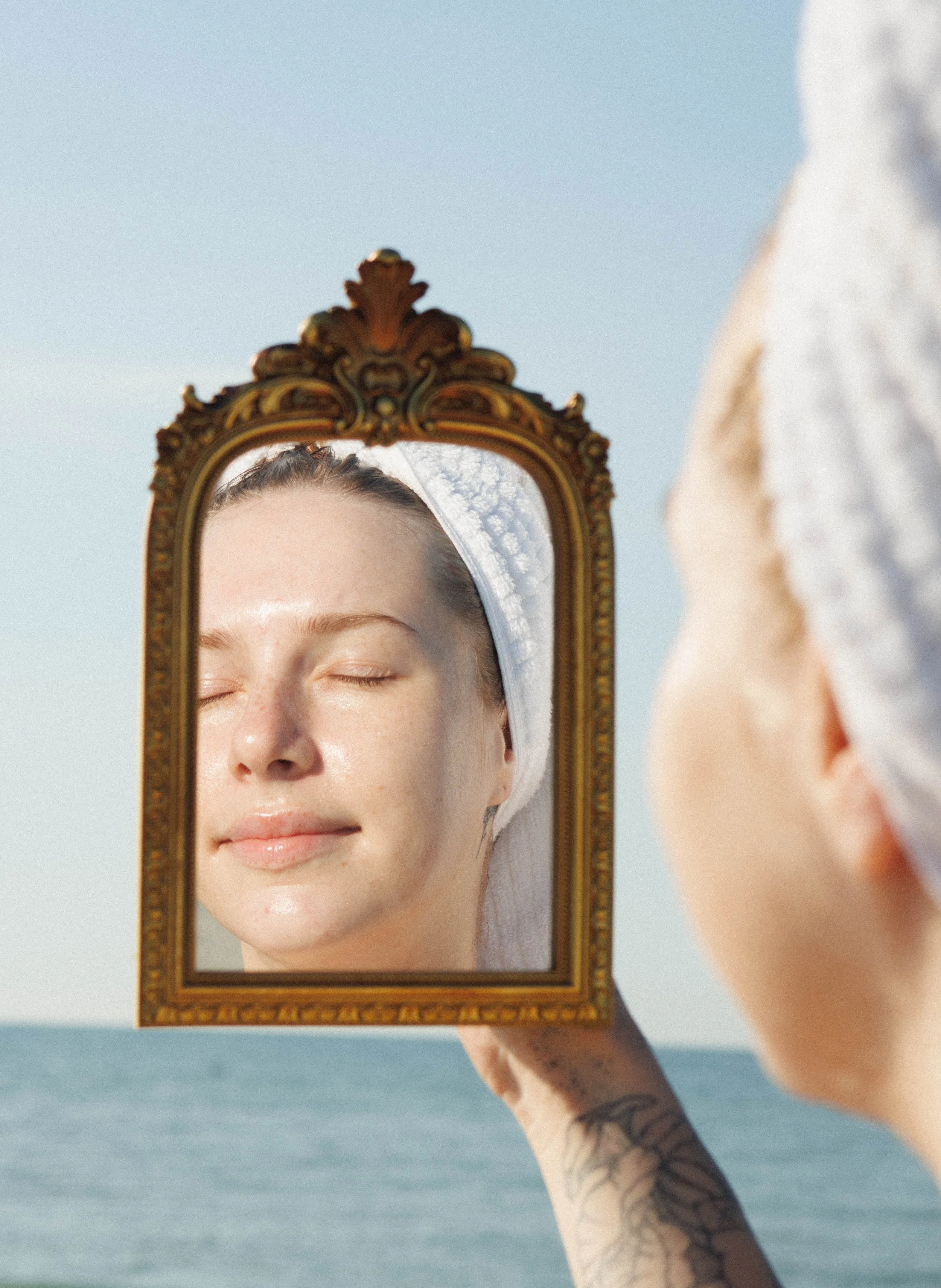 A woman with a towel wrapped around her head is looking at her reflection in an ornate gold-framed mirror she is holding up with one hand, against a background of the ocean and clear sky.
