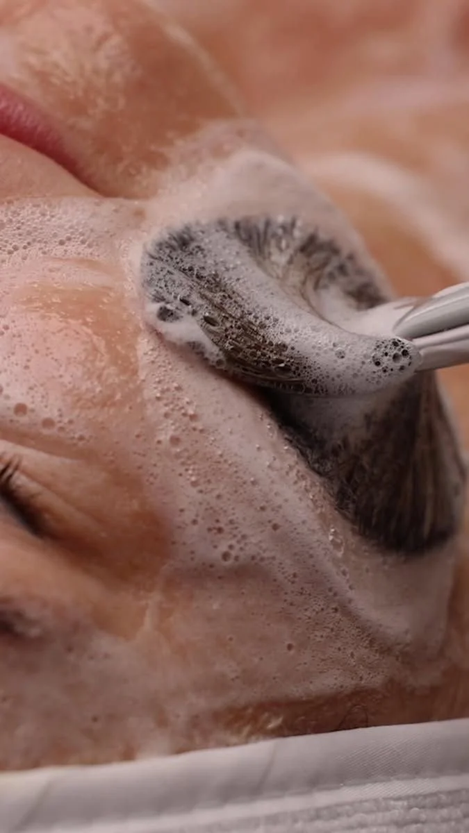 Close-up of a person's face with closed eyes, covered in soap bubbles, receiving a facial cleansing with a brush.
