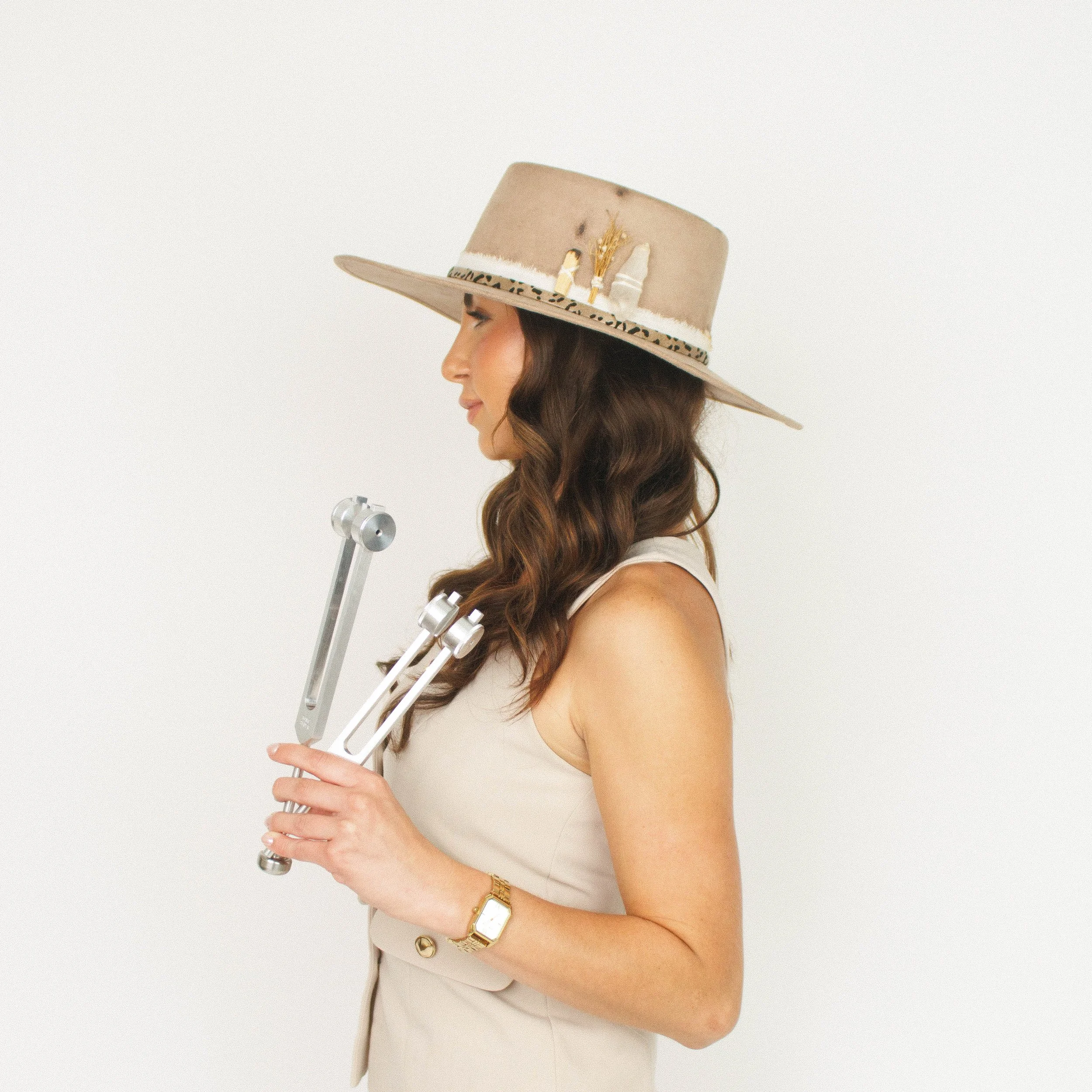 Woman with long wavy brown hair wearing a wide-brim beige hat and beige sleeveless dress, holding a pair of toning chopsticks, standing against a plain white background.