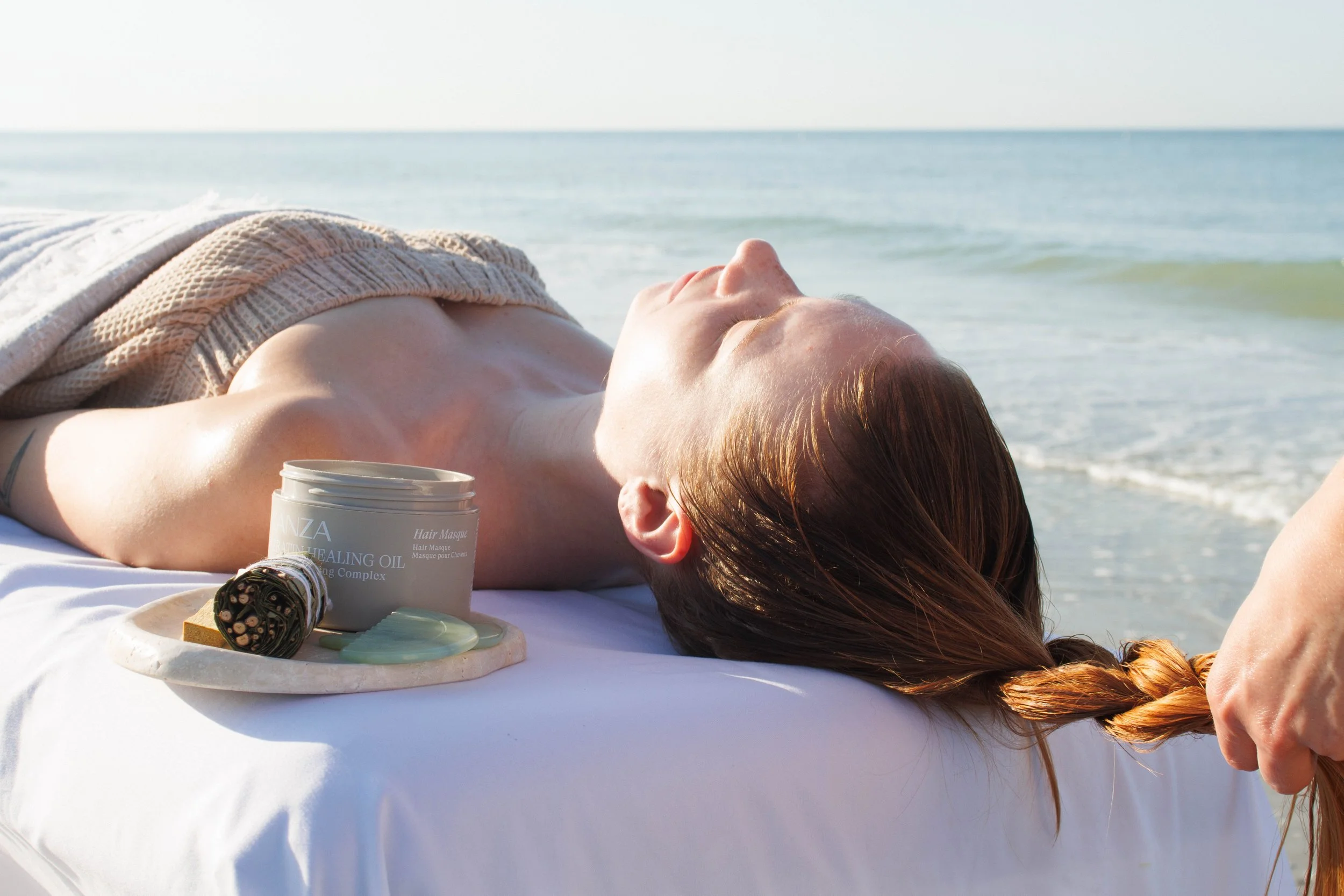 A woman lying on a massage table at the beach, enjoying a relaxing moment with her eyes closed, with ocean waves in the background. Massage oils and skincare products are nearby.