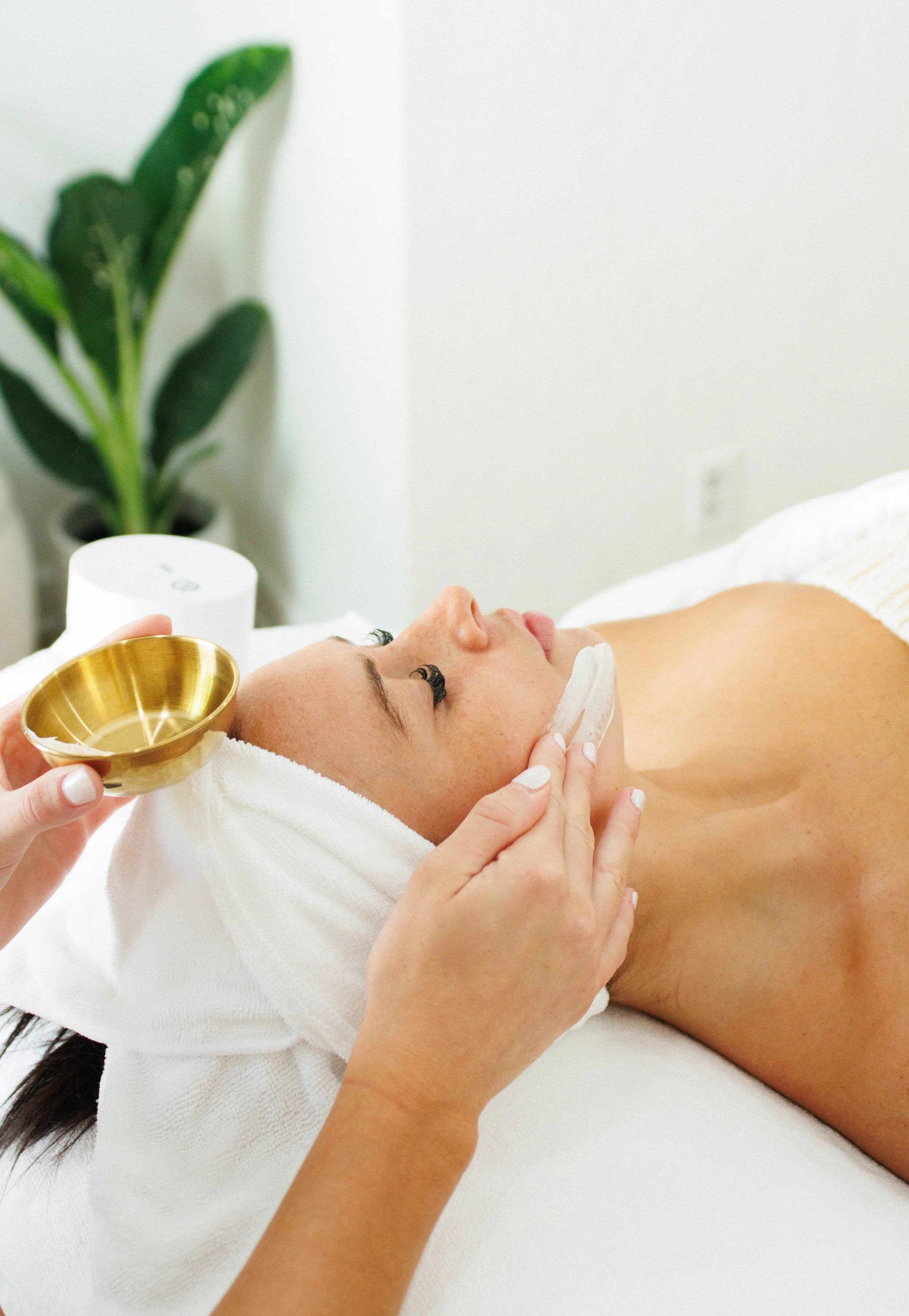 A woman lying on a massage table with a towel wrapped around her head receiving facial treatment, with a person holding a bowl and towel near her face in a spa room.