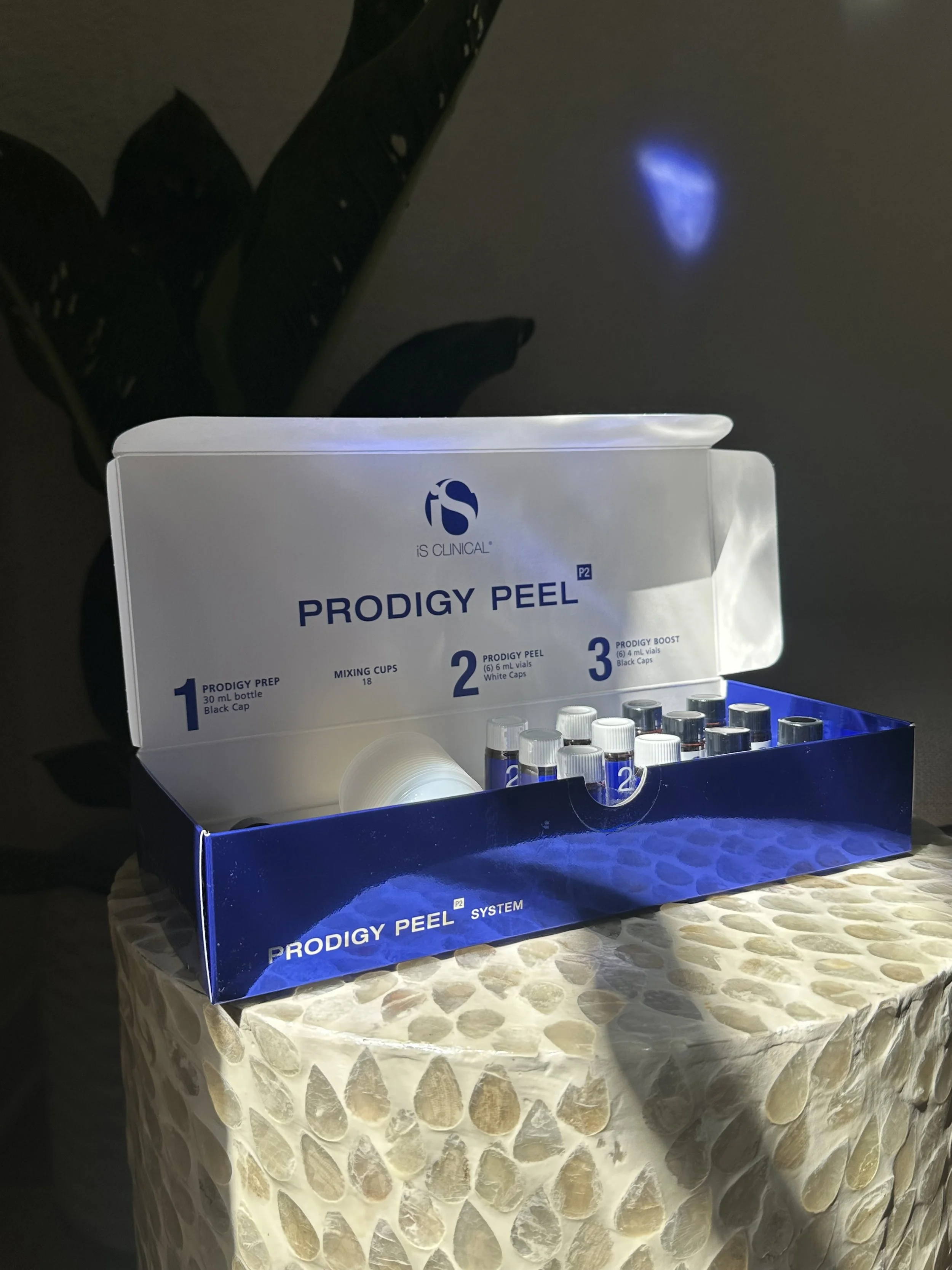 Open box of iS Clinical Prodigy Peel System with vials, on a textured table with a plant in the background. Light is reflecting off the box, creating a glow effect.
