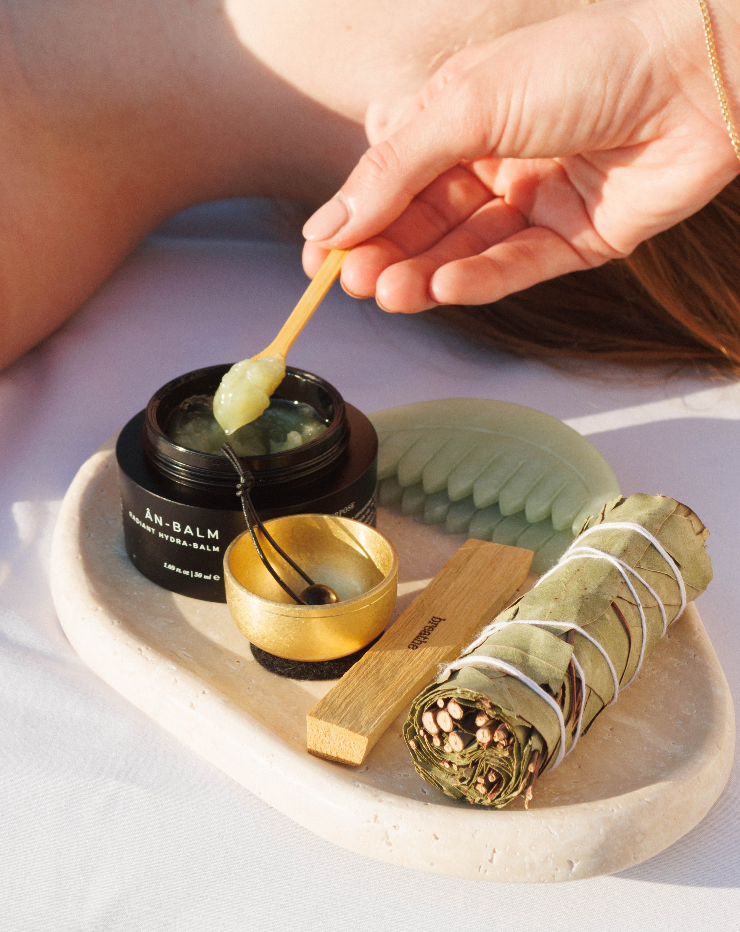 A person applying balm from a black jar labeled 'ÀN-BALM' onto their lips using a wooden spatula. The setup includes a round stone tray with a rolled sage bundle, a wooden stick, a small golden bowl, and a carved green stone, with a white background.