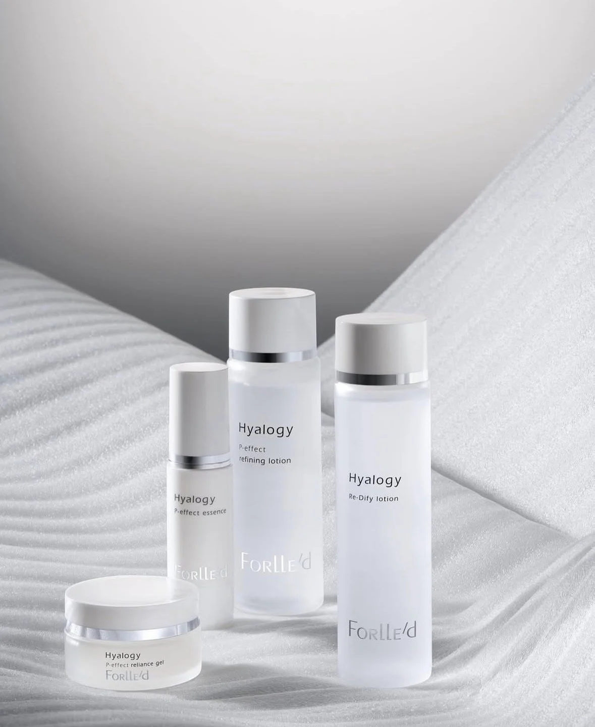 Set of four skincare bottles and jar labeled Hyalogy, including essence, refining lotion, re-dify lotion, and resilience gel, arranged on a textured white surface with white geometric background.