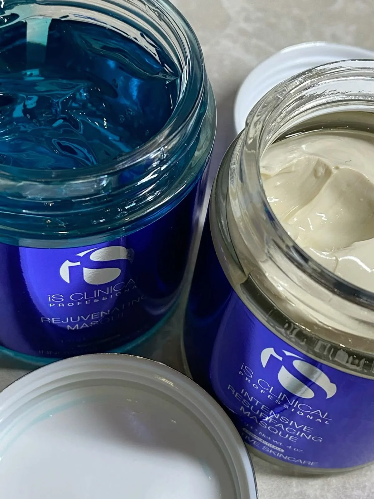 Open jars of skincare products labeled 'Rejuvenating Mask' and 'Intensive Restoring Mask' from iS Clinical, with a closed white lid nearby.