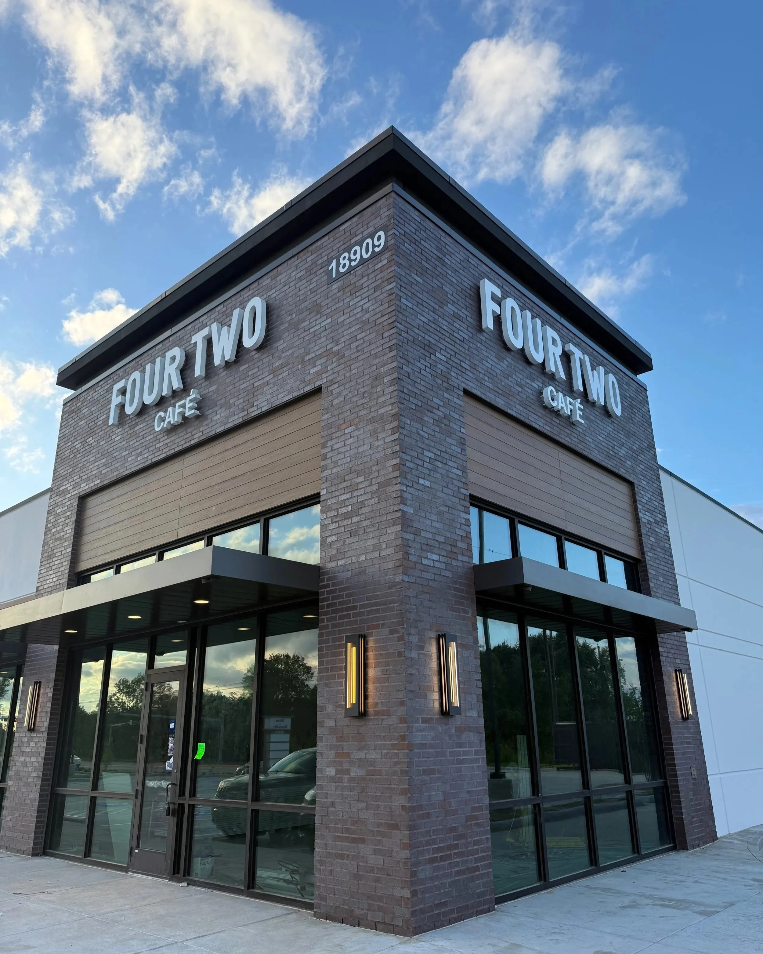 Corner of a modern brick building with large glass windows and signage that reads 'Four Two Cafe' and the address '18909', with a blue sky and white clouds above.