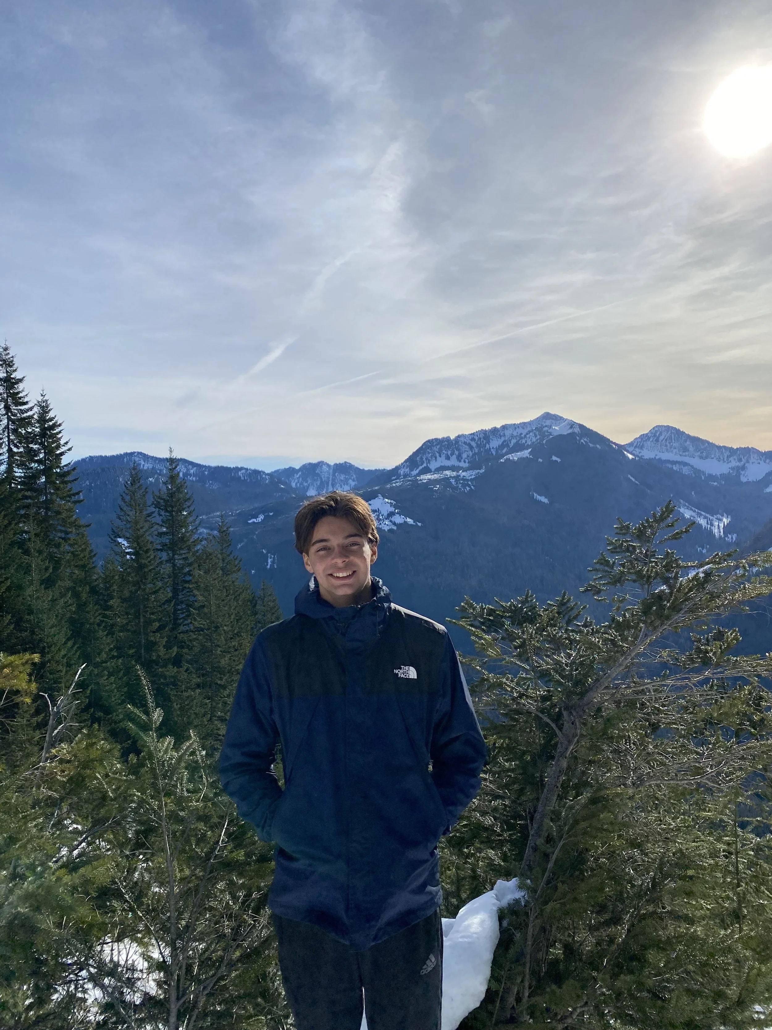 A young man smiling outdoors in front of a mountainous landscape with snow-capped peaks, trees, and a cloudy sky.
