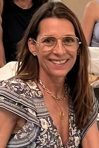 Woman with long brown hair, wearing glasses, a patterned shirt, and gold jewelry, smiling at the camera.