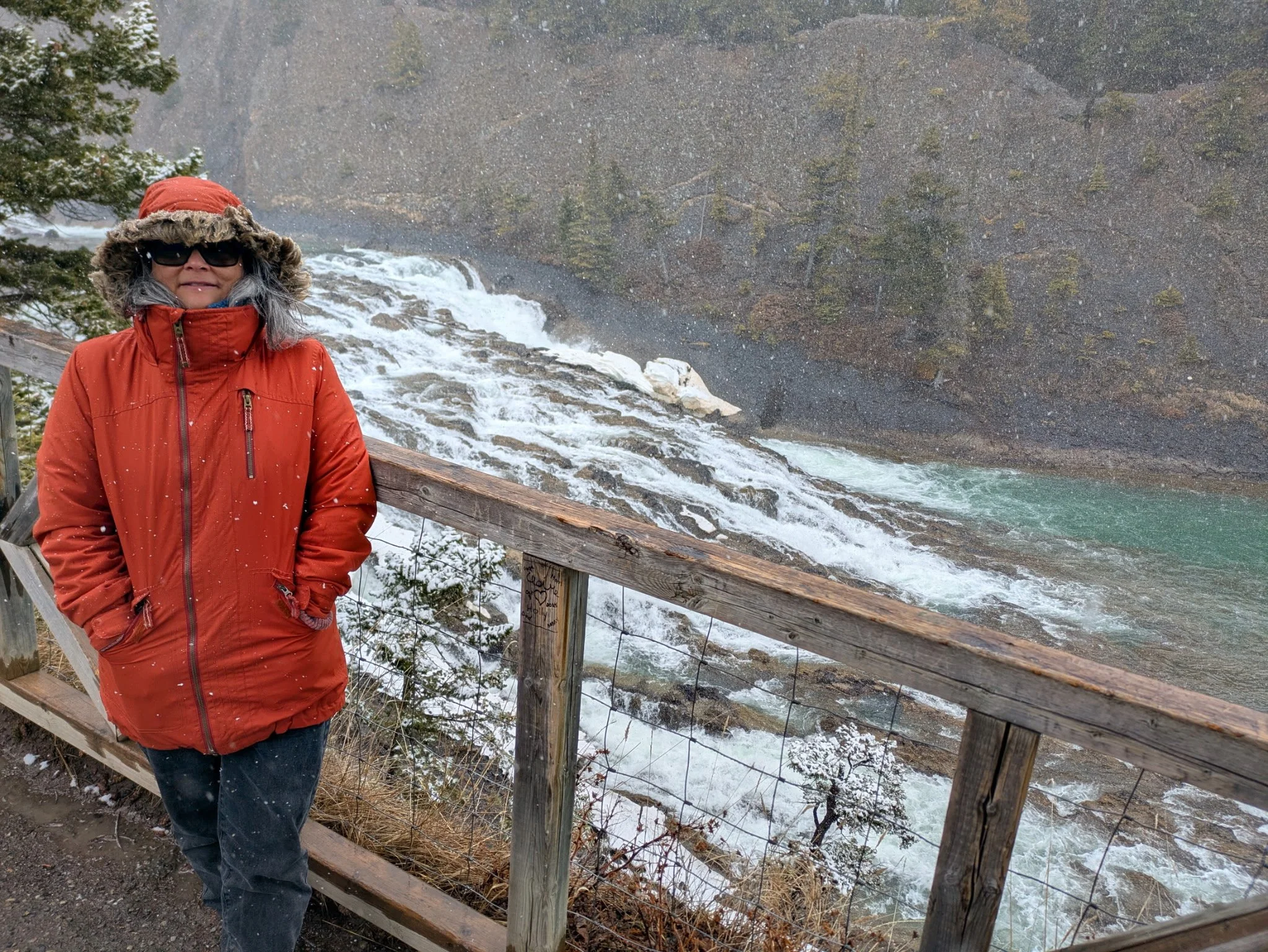 A woman in a red winter jacket and a red hat with fur trim stands near a wooden railing in front of a mountain waterfall. She is wearing sunglasses and has gray hair. Snow is falling and there are snow patches on the ground and on the trees nearby.