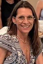 Woman with shoulder-length brown hair, glasses, and a patterned top, smiling at a social gathering.