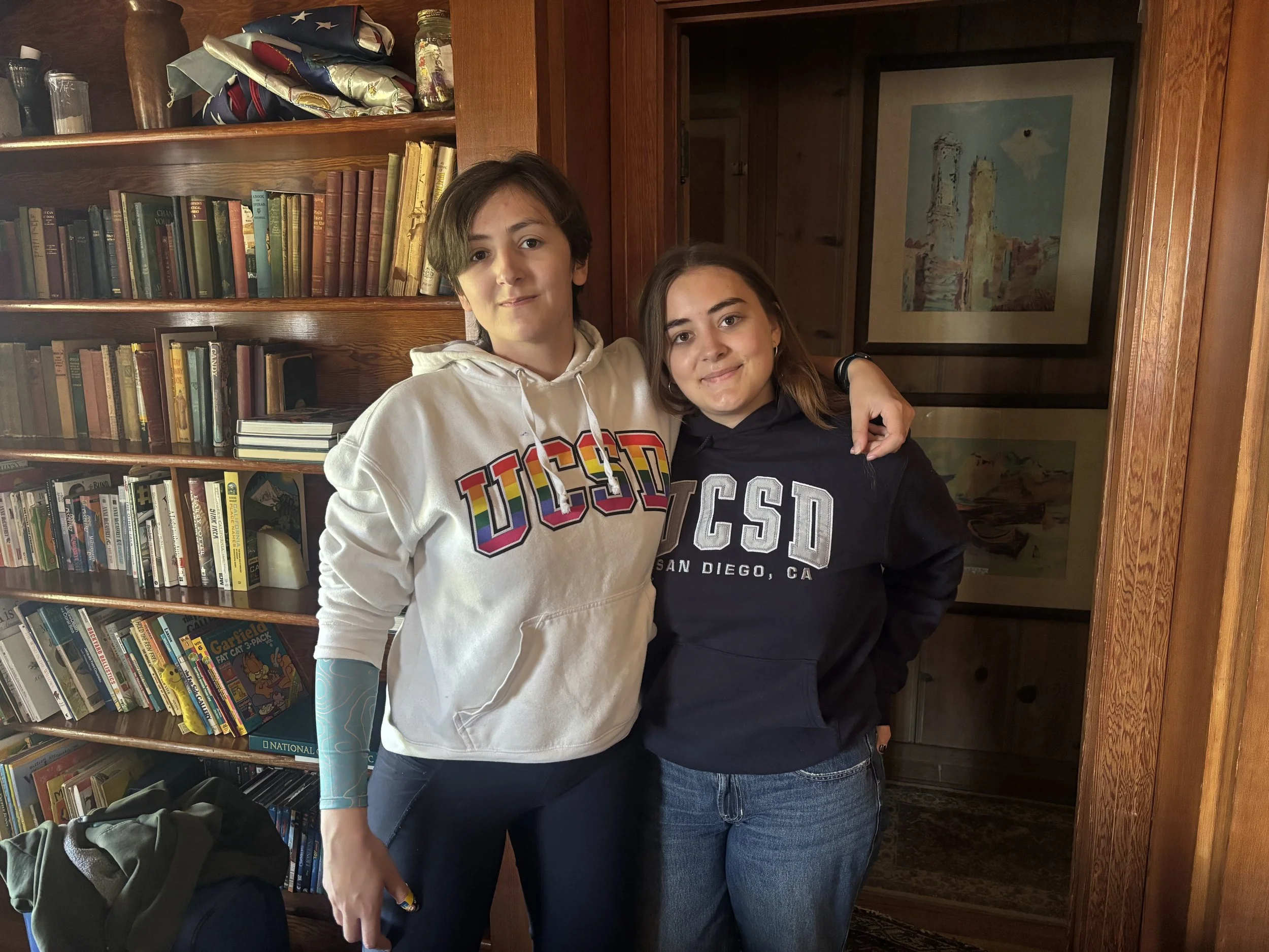 Two young women are standing close together inside a room with wooden bookshelves filled with books behind them. They are smiling and have their arms around each other. One woman is wearing a white hoodie with rainbow-colored text, and the other woman is wearing a black hoodie. The background includes framed artwork on the wall and a small table with a backpack.