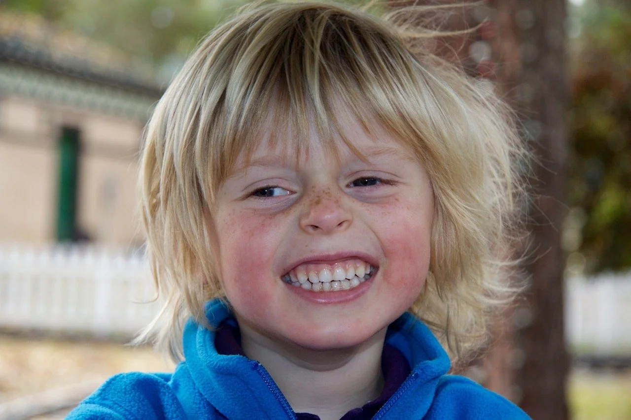 Close-up of a smiling young boy with blond hair, freckles, and chubby cheeks outdoors.