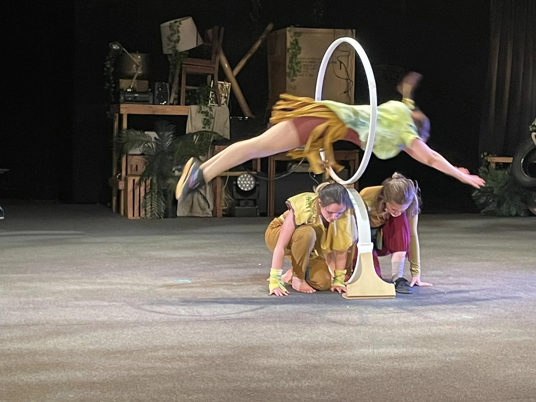 Two children in colorful costumes performing a balancing act on stage, with one child inside a hula hoop and the other bending forward with hands on the floor, in front of a set decorated with plants and furniture.