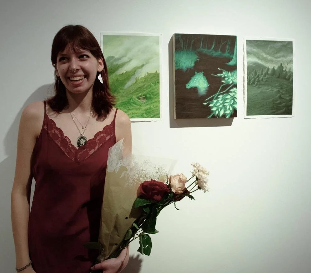A woman with red hair wearing a burgundy dress and jewelry holding a bouquet of roses, standing in front of three abstract landscape paintings on a white wall.