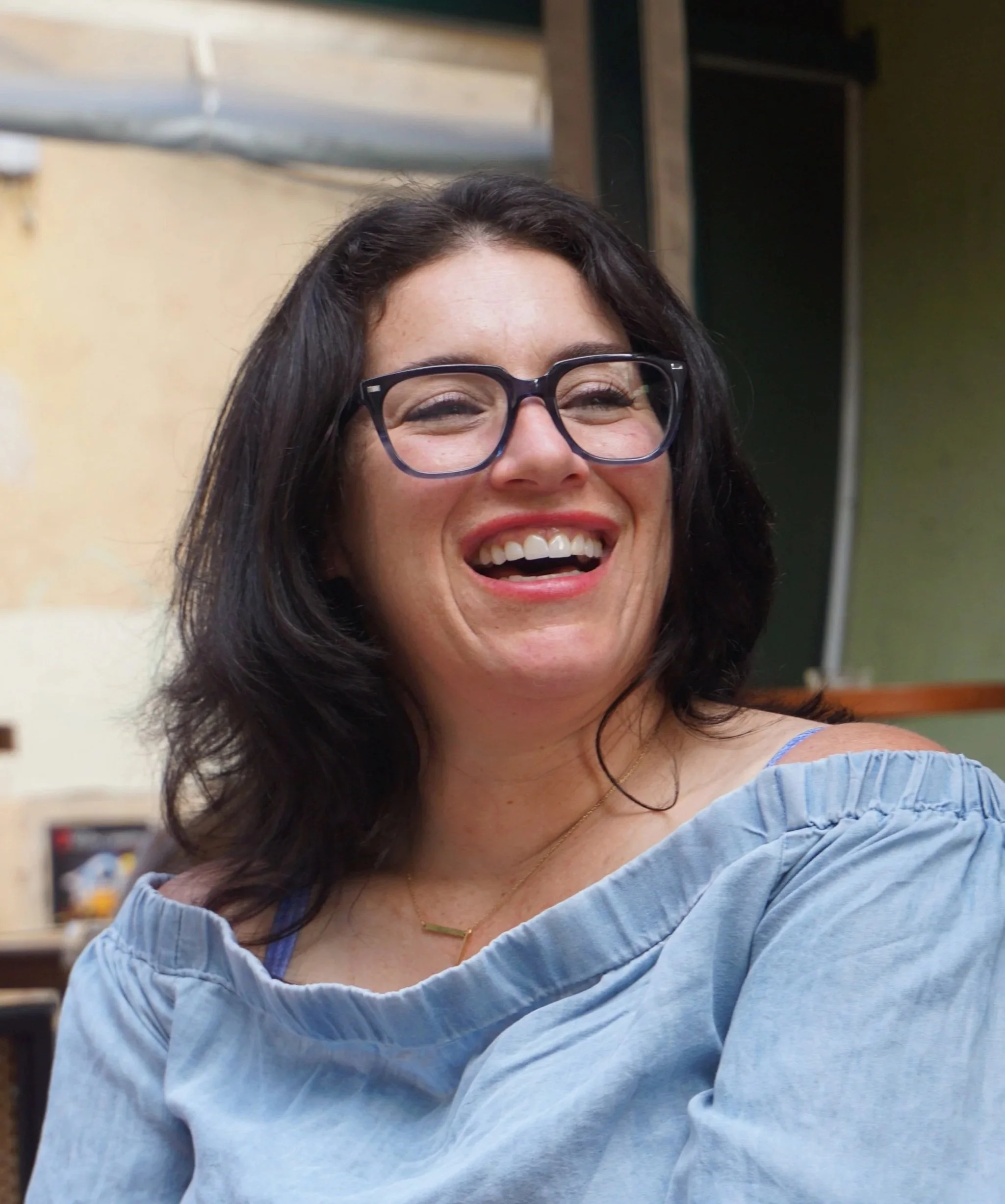Close-up of a woman with dark hair, wearing glasses and a light blue off-the-shoulder top, smiling in an indoor setting.