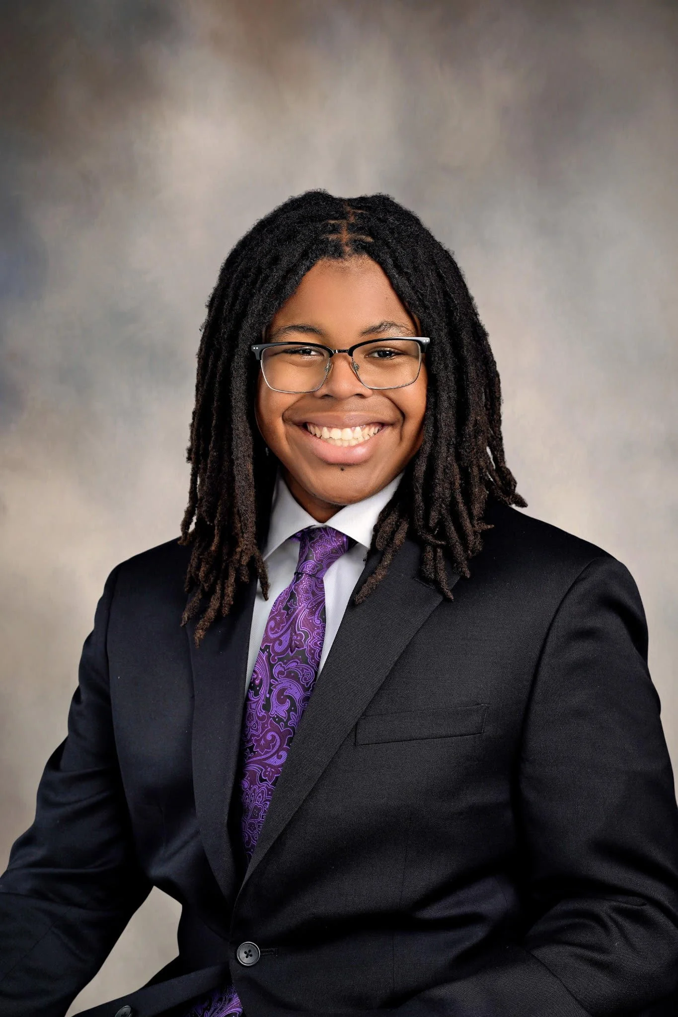 Portrait of a smiling woman with glasses, dressed in a black suit, white shirt, and purple tie, against a neutral background.