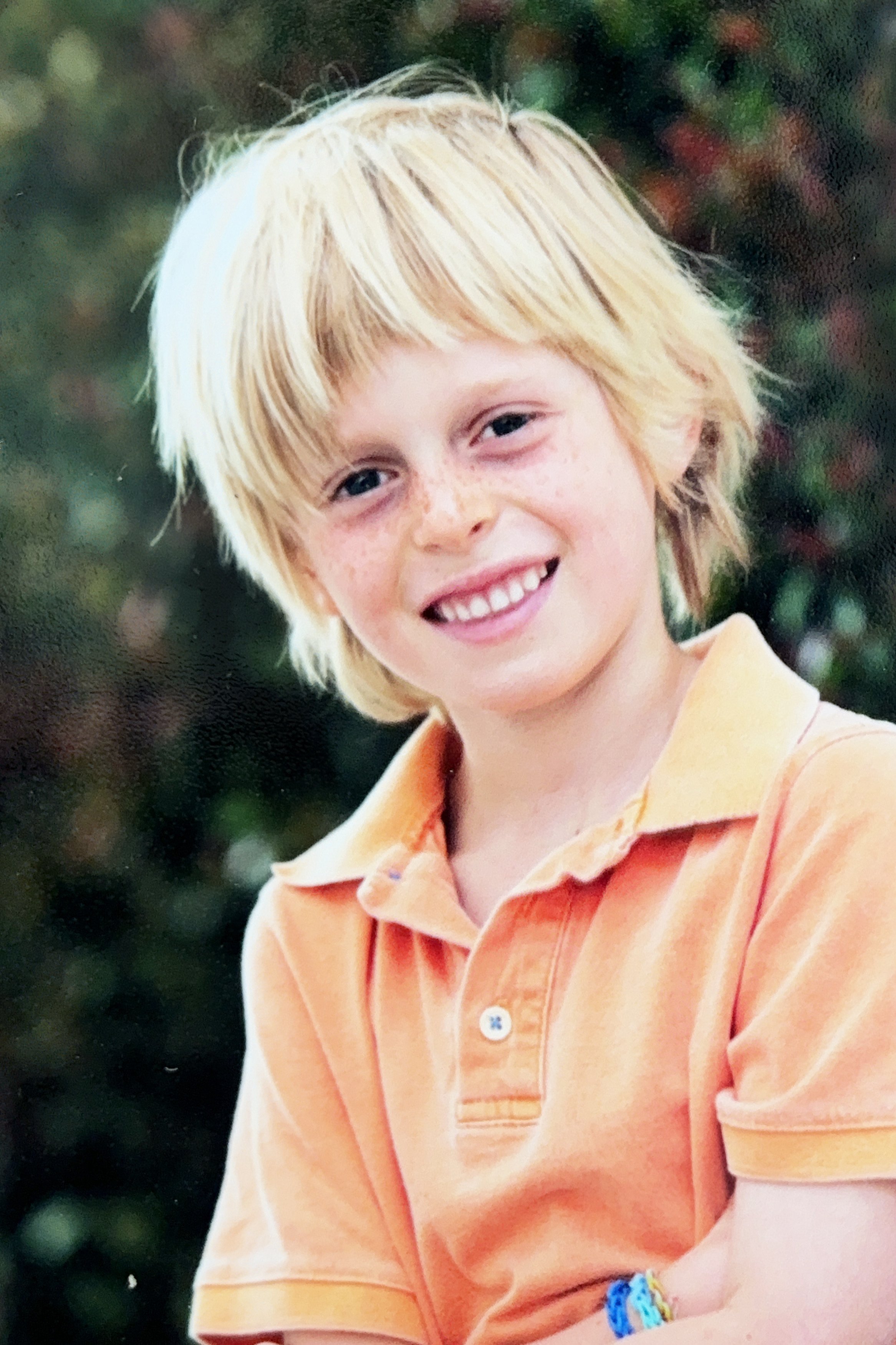 A young boy with blonde hair and freckles smiling, wearing an orange collared shirt, with a background of blurred greenery.
