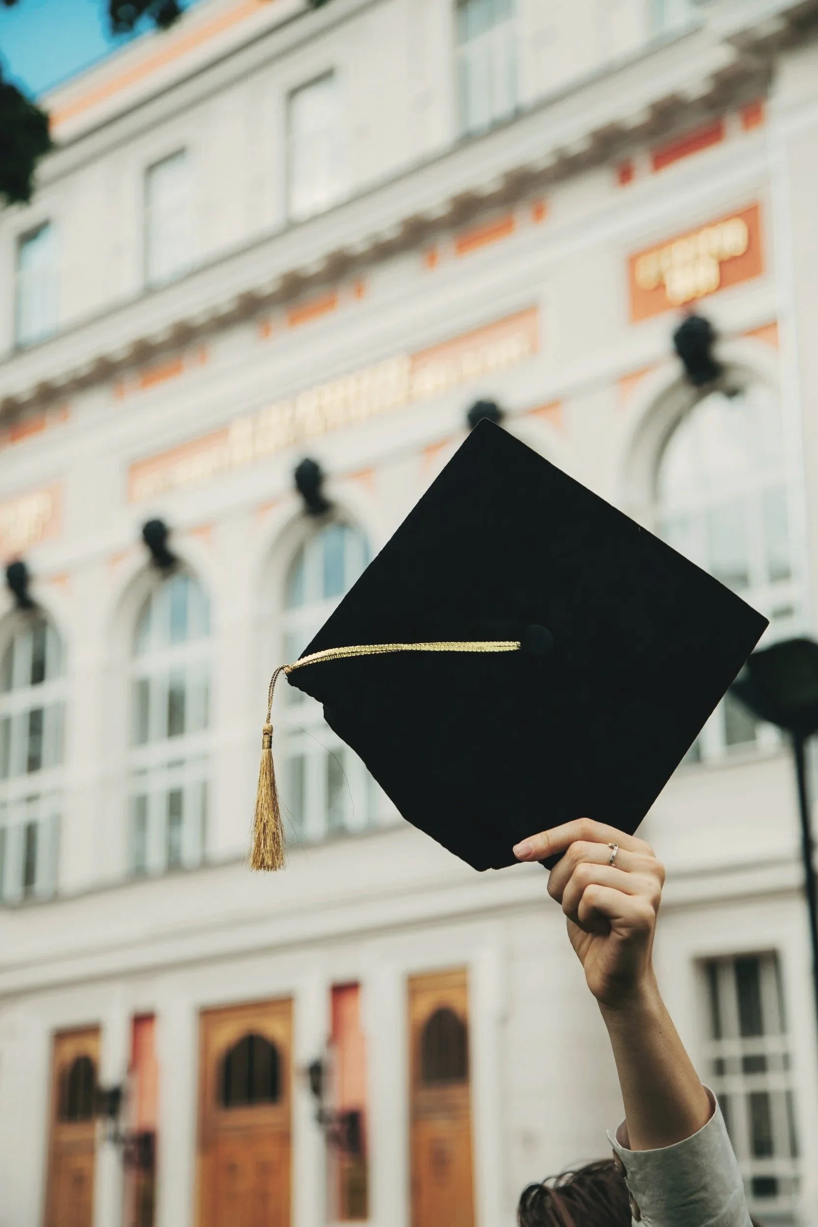 A person holding a black graduation cap with a gold tassel in front of a historic building with large windows and decorative architectural elements.