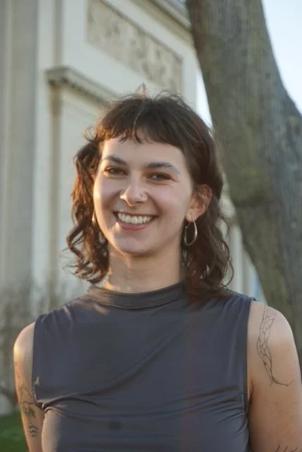 A young woman with short, curly brown hair and hoop earrings, smiling outdoors with a large tree and building in the background.