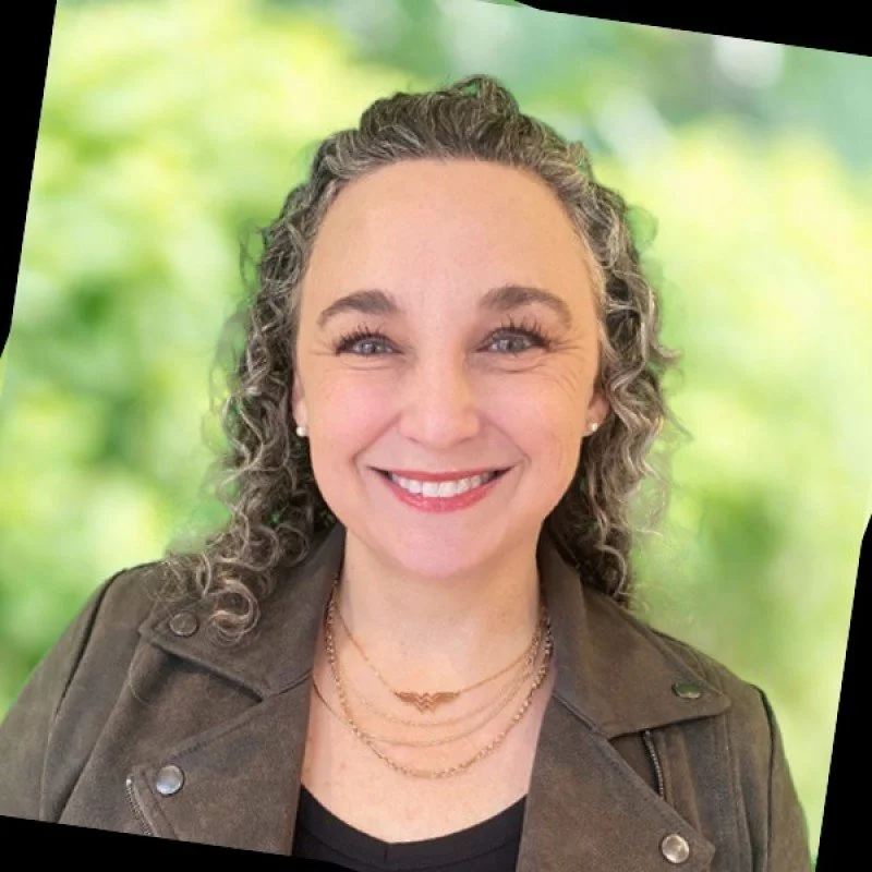 A smiling woman with curly gray hair, wearing a brown leather jacket and layered necklaces, against a blurred green outdoor background.